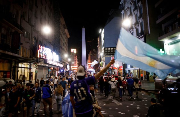Rua noturna em Buenos Aires com o Obelisco. Multidão celebra, um torcedor com camisa do Messi ergue bandeira argentina.