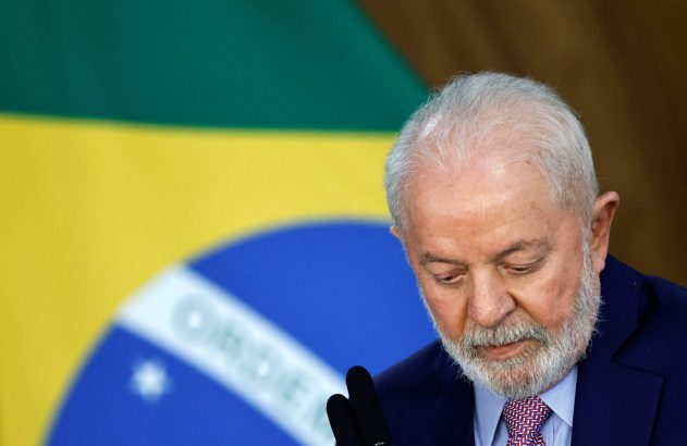 Presidente Lula da Silva, de terno, olhando para baixo, com a bandeira do Brasil ao fundo.