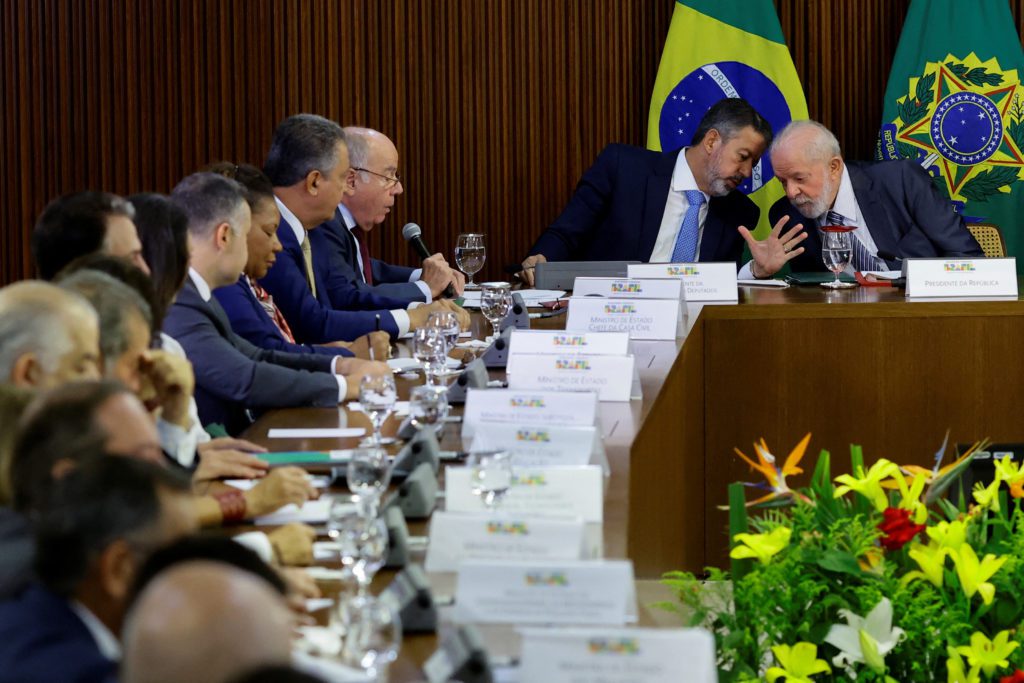 Lula conversa de perto com um homem em reunião governamental. Várias autoridades estão sentadas à mesa.