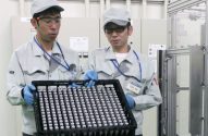 Dois técnicos uniformizados seguram bandeja de células de bateria em ambiente industrial.