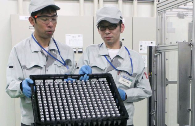 Dois técnicos uniformizados seguram bandeja de células de bateria em ambiente industrial.