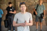 Sam Altman está sorrindo para a câmera, de camiseta clara e óculos na cabeça. Pessoas e parede de pedra ao fundo
