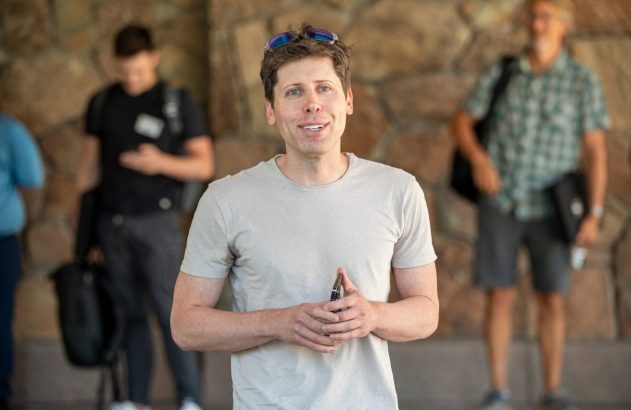 Sam Altman está sorrindo para a câmera, de camiseta clara e óculos na cabeça. Pessoas e parede de pedra ao fundo