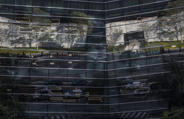 Fachada de prédio de vidro refletindo árvores, carros na rua e pessoas.