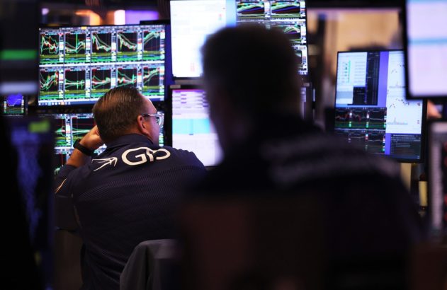 Pessoa de costas em sala de negociações, observando monitores com gráficos financeiros. Outra pessoa desfocada em primeiro plano.