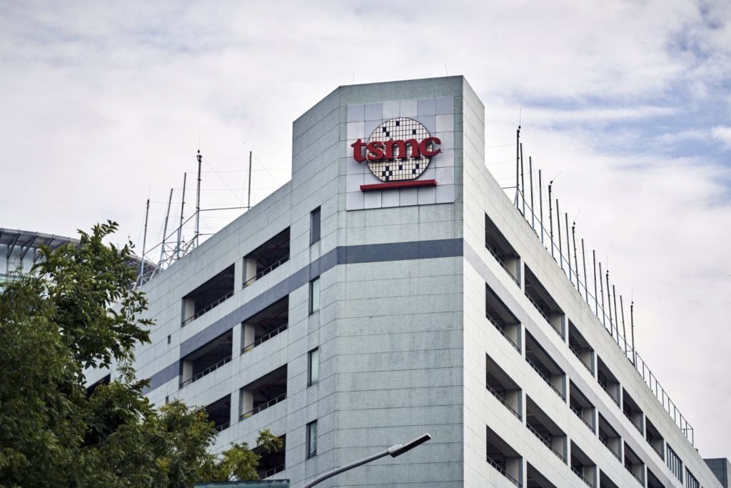 Vista externa do prédio da TSMC com seu logo vermelho sobre um design de wafer. O edifício tem fachada cinza e antenas no telhado.