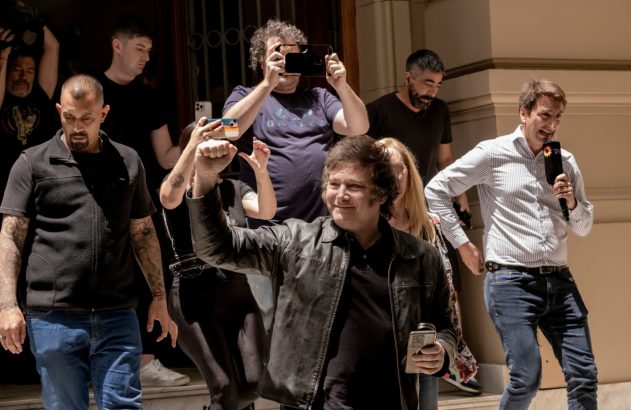 Homem de jaqueta preta sorri e ergue o punho, cercado por apoiadores e um repórter com microfone.