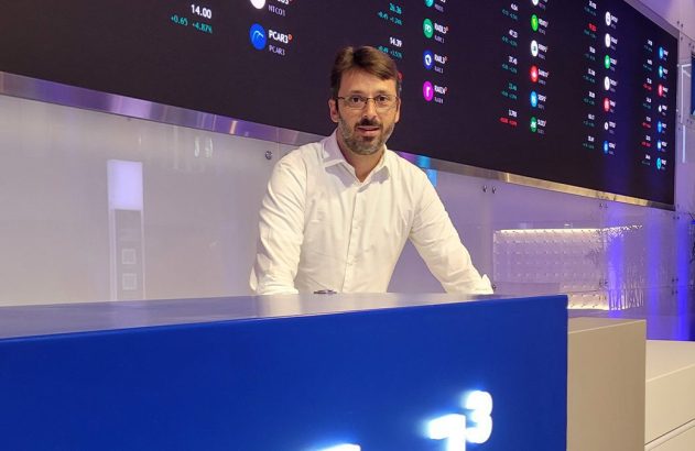 Homem de camisa branca em balcão azul. Ao fundo, tela digital mostra cotações da B3.