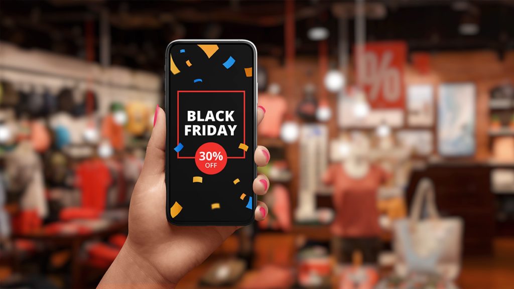 Mão segurando smartphone com promoção de Black Friday (30% off) na tela, fundo de loja desfocado.