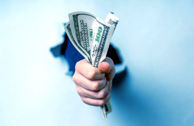 Mão de uma pessoa segurando notas de 100 dólares, emergindo de um rasgo em uma superfície azul.