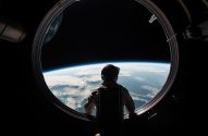 Pessoa de costas observa a Terra com nuvens e o espaço através de uma escotilha circular de nave espacial.