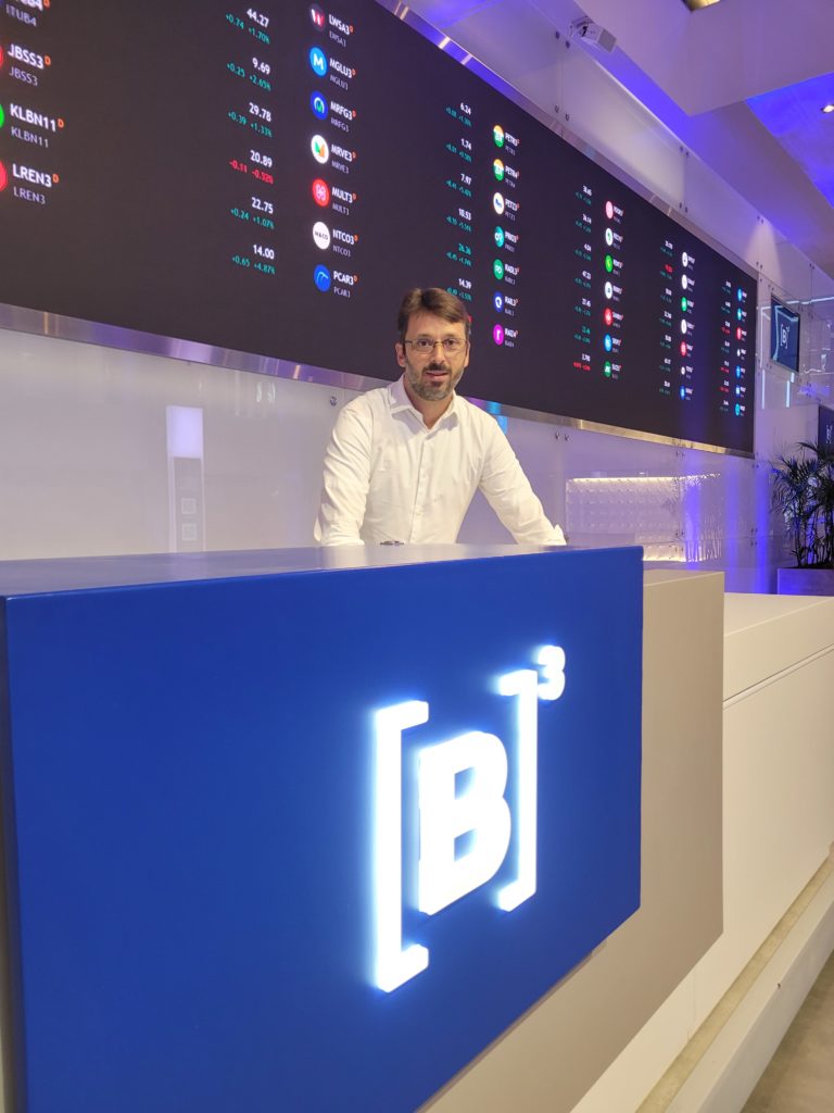 Um homem sorrindo atrás de um balcão azul com o logo B3, e tela gigante de cotações da bolsa ao fundo.