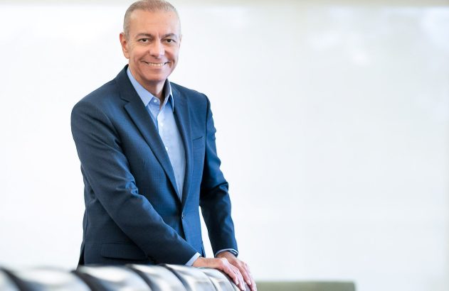 Homem sorridente de terno azul com as mãos no encosto, fundo branco.