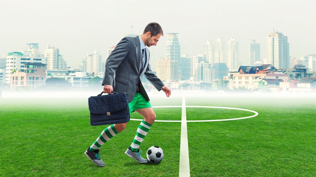 Executivo com traje de futebol e maleta chuta bola em campo. Cidade ao fundo.