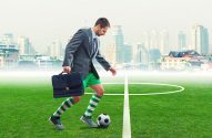 Executivo com traje de futebol e maleta chuta bola em campo. Cidade ao fundo.