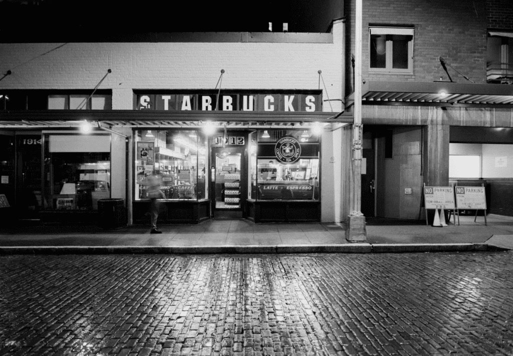 Fachada noturna em preto e branco da primeira Starbucks, com rua de paralelepípedos molhada.