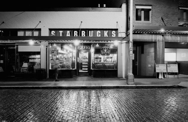 Fachada noturna em preto e branco da primeira Starbucks, com rua de paralelepípedos molhada.