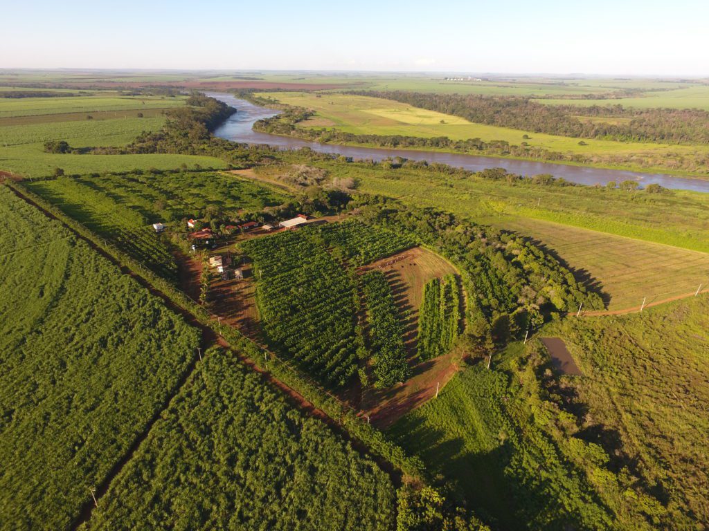 Vista aérea de fazenda e plantações extensas à beira de um rio sinuoso em paisagem rural ensolarada.