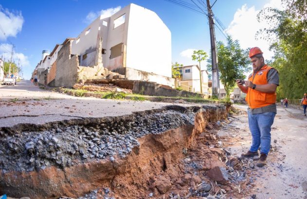 Homem em colete laranja e capacete inspeciona uma rua com asfalto quebrado e erosão severa, com prédios danificados ao fundo.