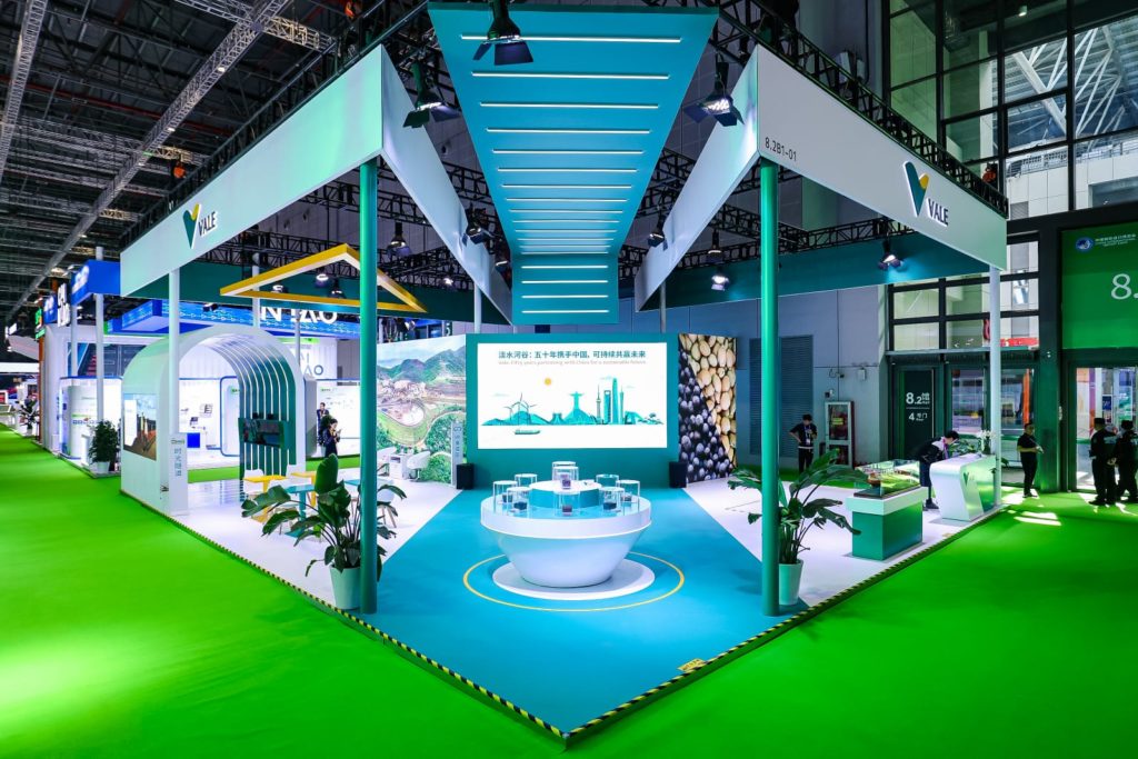 Estande da Vale em feira: design moderno verde e azul, telas digitais, balcão de amostras e visitantes.