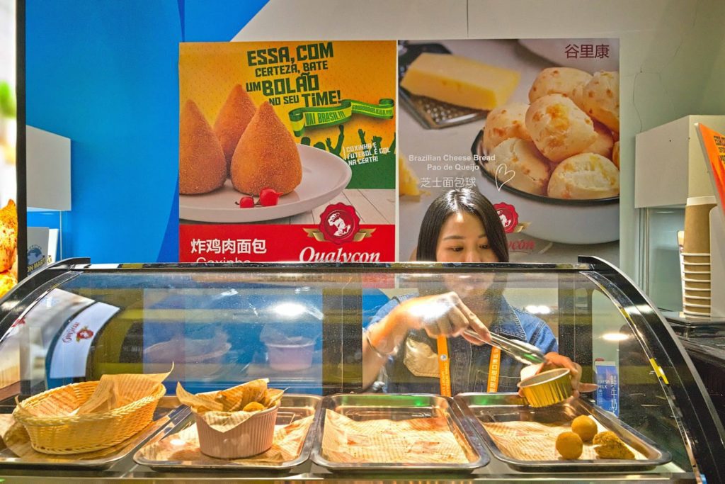 Mulher servindo salgados brasileiros (coxinha e pão de queijo) em balcão de atendimento.