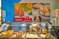 Mulher servindo salgados brasileiros (coxinha e pão de queijo) em balcão de atendimento.