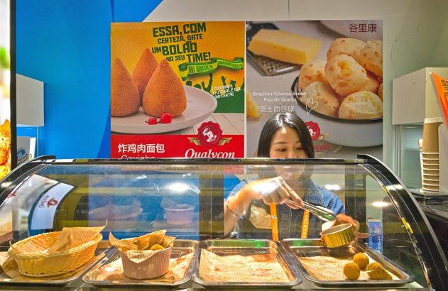 Mulher servindo salgados brasileiros (coxinha e pão de queijo) em balcão de atendimento.