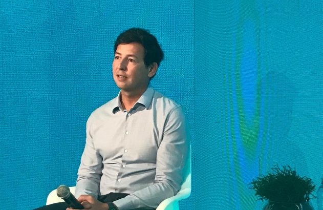 Homem sentado de camisa cinza clara segura um microfone e olha para a esquerda em frente a um fundo azul.