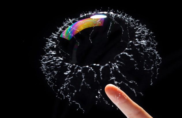 Dedo toca bolha colorida, que estoura em respingos de água formando um anel. Fundo preto.