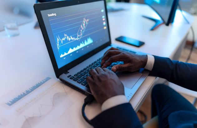 mãos negras sobre teclado de notebook com gráficos financiros na tela do computador.