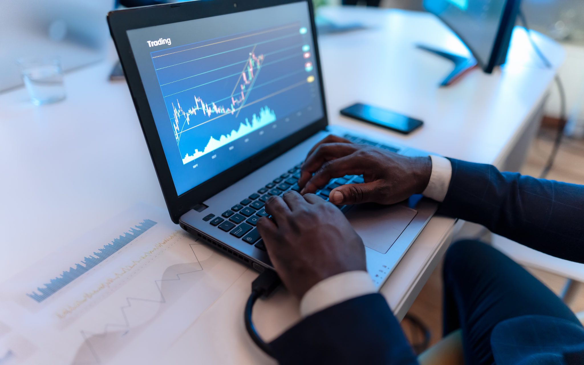 mãos negras sobre teclado de notebook com gráficos financiros na tela do computador.