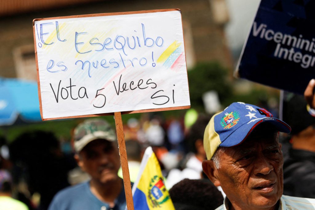 Cartaz em manifestação: 'El Esequibo es nuestro! Vota 5 Veces Si'. Homem com boné da Venezuela.