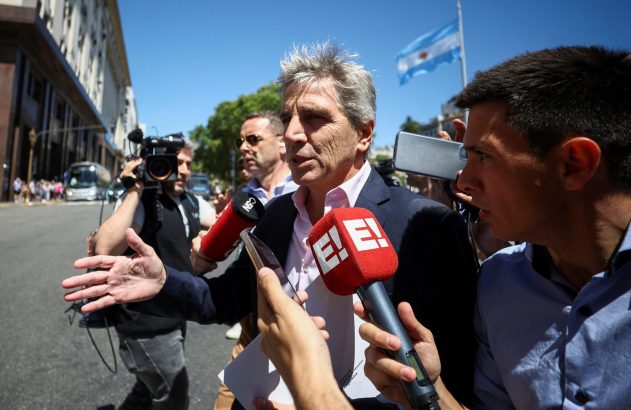 Homem de terno grisalho fala a jornalistas com microfones e celulares em coletiva de imprensa ao ar livre. Bandeira argentina ao fundo.