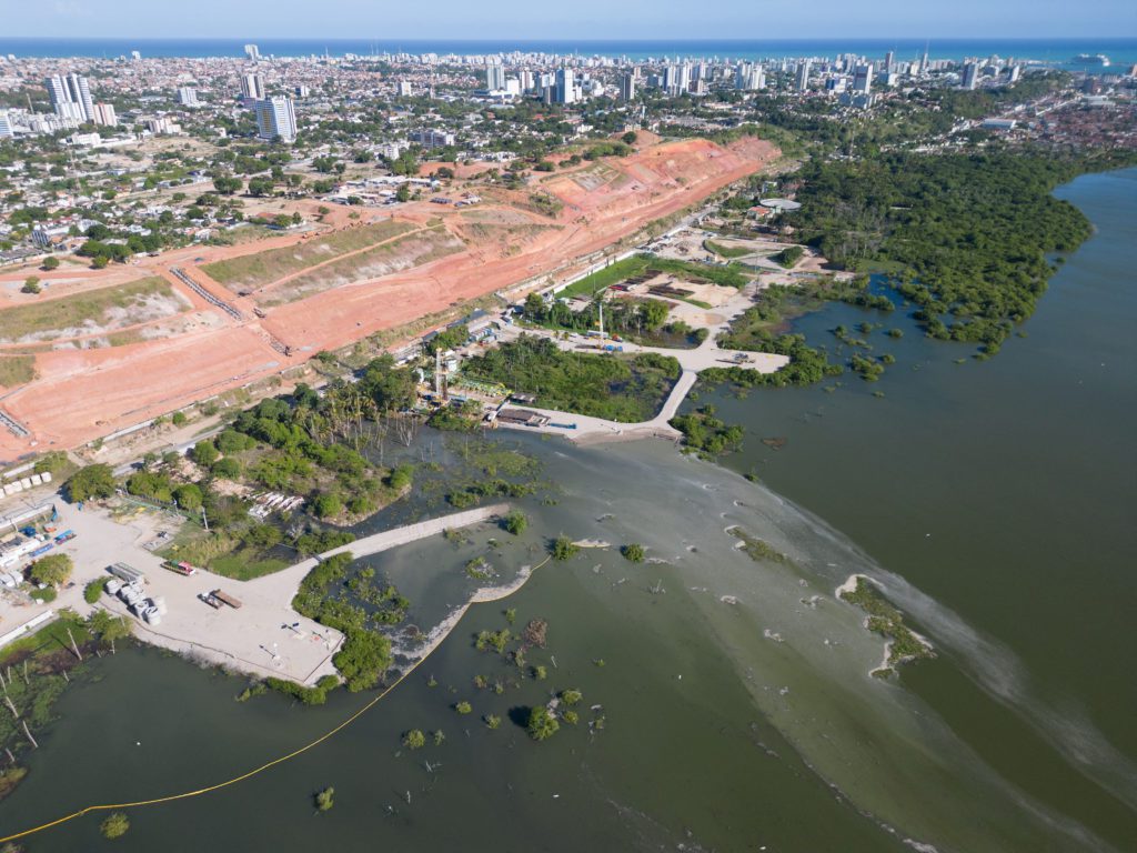 Vista aérea: cidade costeira, canteiro de obras em terra vermelha e estuário com água verde-turva.