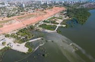 Vista aérea: cidade costeira, canteiro de obras em terra vermelha e estuário com água verde-turva.