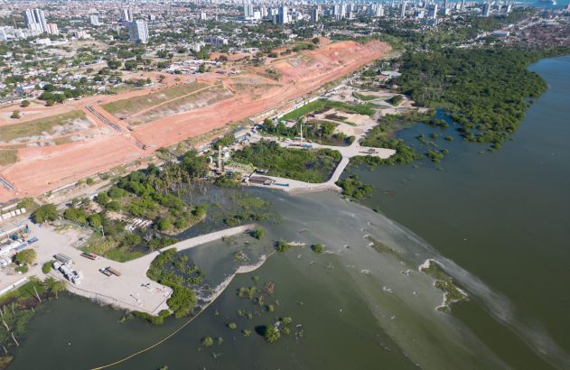 Vista aérea: cidade costeira, canteiro de obras em terra vermelha e estuário com água verde-turva.