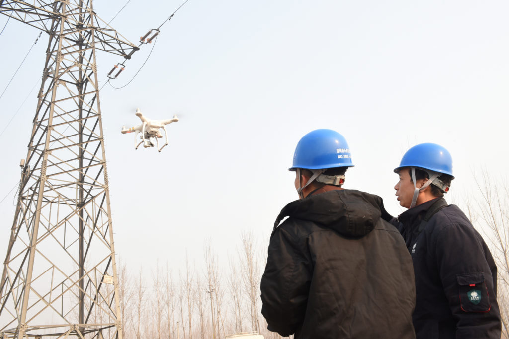 Dois técnicos com capacete azul observam um drone inspecionando uma torre de energia.