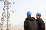 Dois técnicos de capacete azul observam drone inspecionando torre de transmissão de energia.