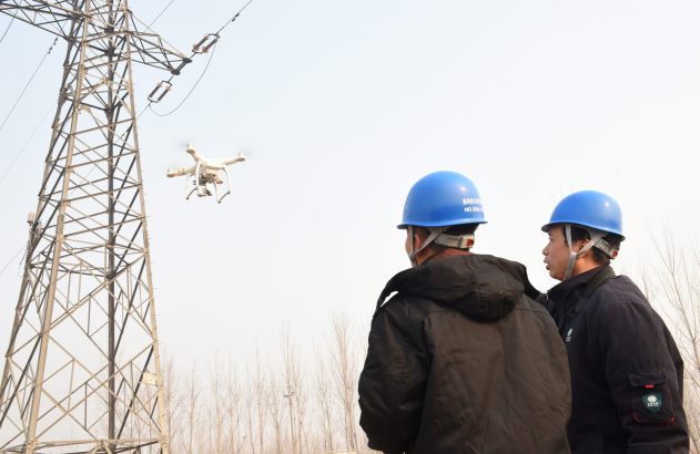 Dois técnicos de capacete azul observam drone inspecionando torre de transmissão de energia.