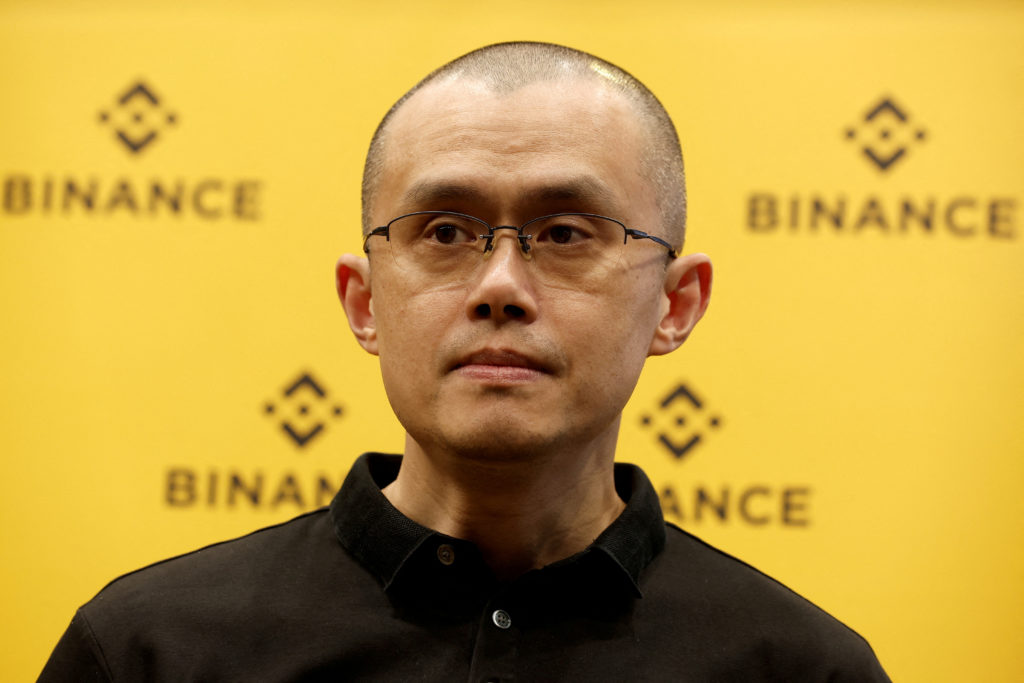 Changpeng Zhao, CEO da Binance, de óculos e camisa preta, contra fundo amarelo com logo Binance.