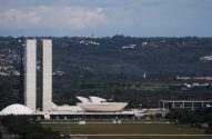 Vista panorâmica do Congresso Nacional, Brasília: duas torres altas e plenários, sob céu nublado e morros arborizados.