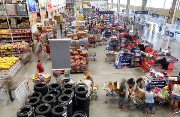 Vista de cima de um grande mercado atacadista. Muitos clientes, produtos nas prateleiras e caixas de atendimento. Pessoas em movimento.