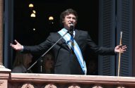 Homem de terno e faixa presidencial argentina discursa em balcão, segurando um bastão e com braços abertos.