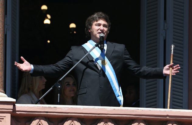 Homem de terno e faixa presidencial argentina discursa em balcão, segurando um bastão e com braços abertos.