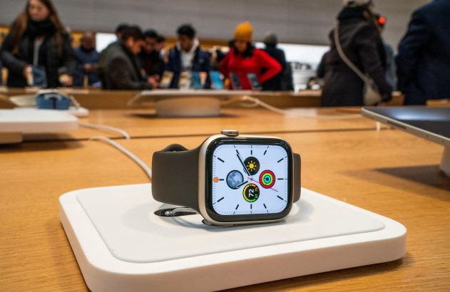 Apple Watch em display sobre base branca em mesa de madeira, com pessoas desfocadas em loja.