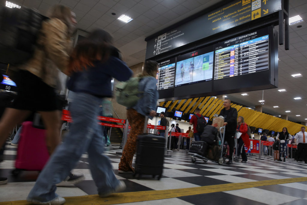 Pessoas com bagagens passam por painel de partidas em aeroporto movimentado. Chão xadrez preto e branco.