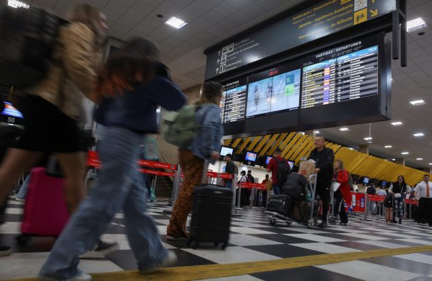 Pessoas com bagagens passam por painel de partidas em aeroporto movimentado. Chão xadrez preto e branco.
