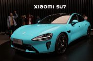 Carro Xiaomi SU7 azul em exibição, com pessoas tirando fotos ao fundo e letreiro "Xiaomi SU7".
