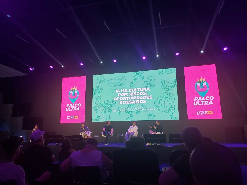 Palco de evento com pessoas no palco e três telas grandes (duas rosas, uma verde). Plateia assiste.