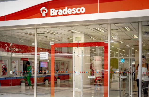 Entrada de agência bancária Bradesco de vidro. Pessoas visíveis, uma usando caixa eletrônico.